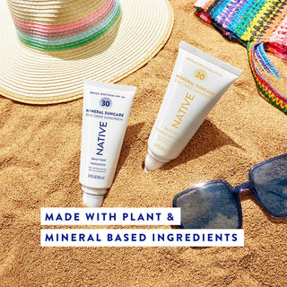 Two Native Mineral Suncare tubes in sand next to sunglasses and a sun hat, labeled plant and mineral-based ingredients